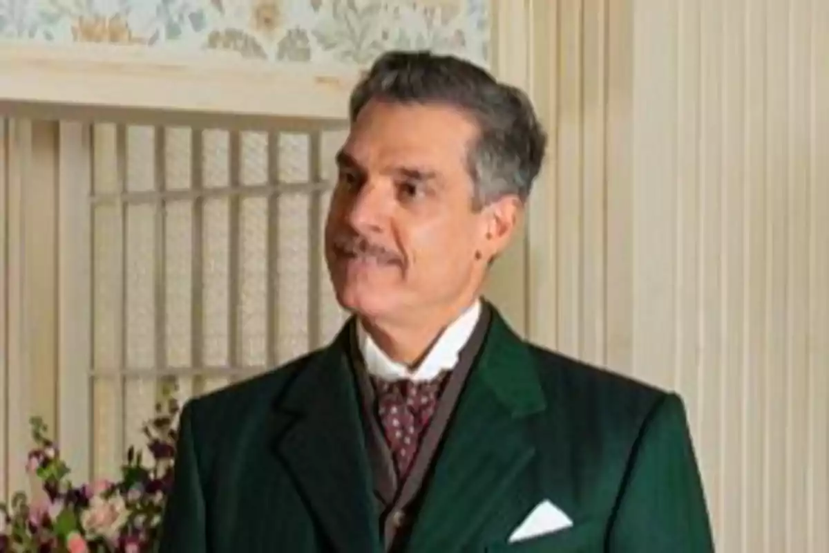 Lisandro amb bigoti i cabell canós porta un vestit verd fosc i corbata, dret davant d'una paret decorada amb paper pintat floral. Captura de 'La Promesa' Lisandro amb bigoti i cabell canós porta un vestit verd fosc i corbata, dret davant d'una paret decorada amb paper pintat floral. Captura de 'La Promesa'
