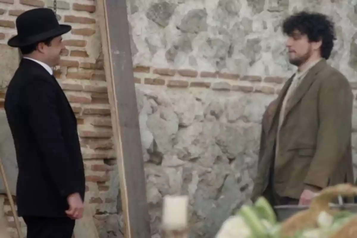 Manuel i Toño conversen davant d'una paret de pedra, un porta un vestit negre i barret, l'altre porta un vestit marró. Captura de 'La Promesa' Manuel i Toño conversen davant d'una paret de pedra, un porta un vestit negre i barret, l'altre porta un vestit marró. Captura de 'La Promesa'