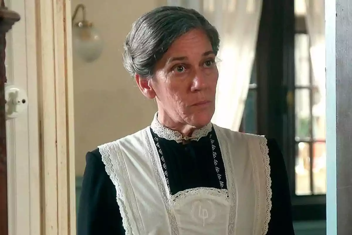 Petra amb els cabells canosos recollits, vestida amb un uniforme de minyona antic en una casa elegant. Captura de 'La Promesa' Petra amb els cabells canosos recollits, vestida amb un uniforme de minyona antic en una casa elegant. Captura de 'La Promesa'