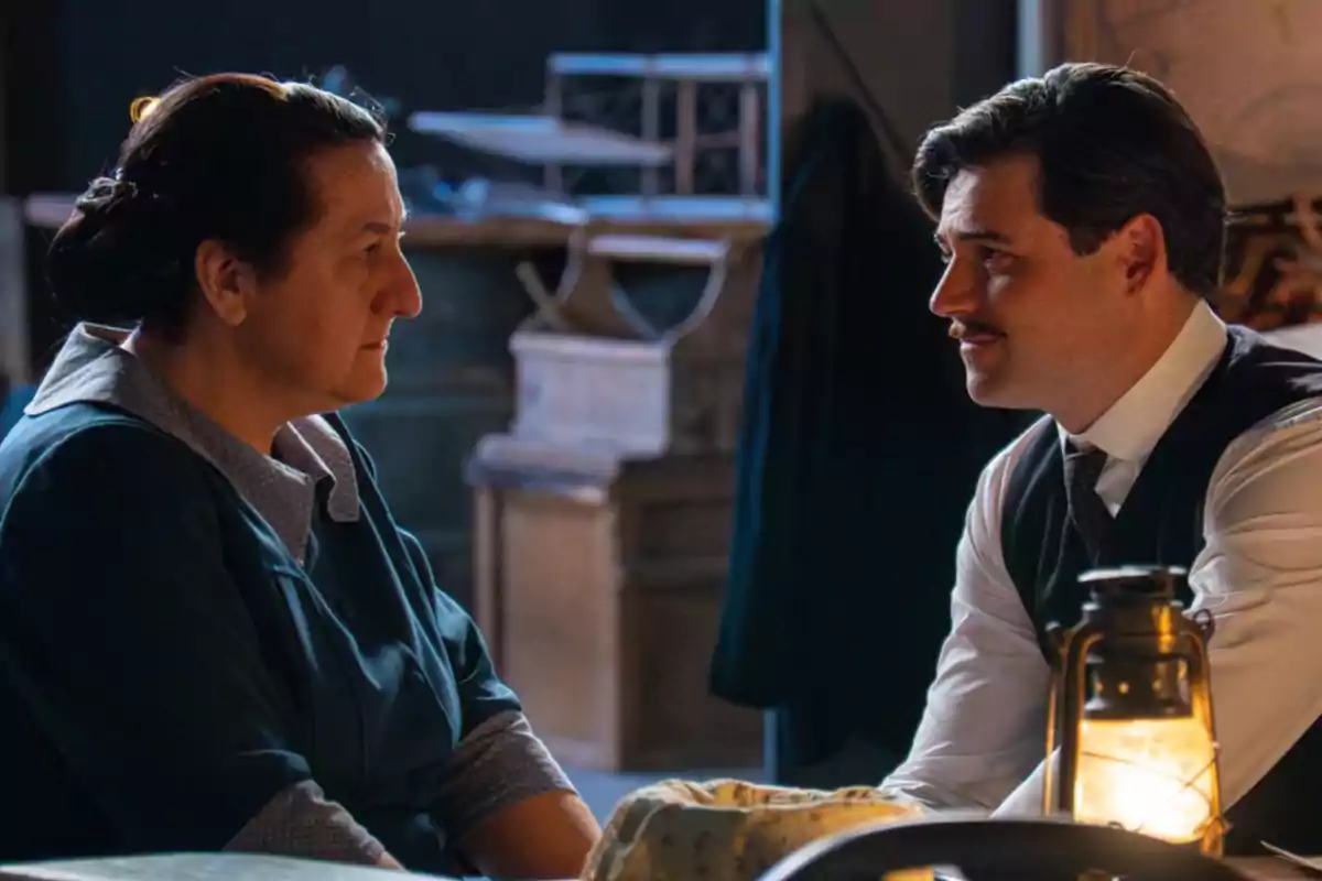 La Simona i en Manuel es miren asseguts cara a cara en una habitació il·luminada per una làmpada antiga. Captura de 'La Promesa' La Simona i en Manuel es miren asseguts cara a cara en una habitació il·luminada per una làmpada antiga. Captura de 'La Promesa'
