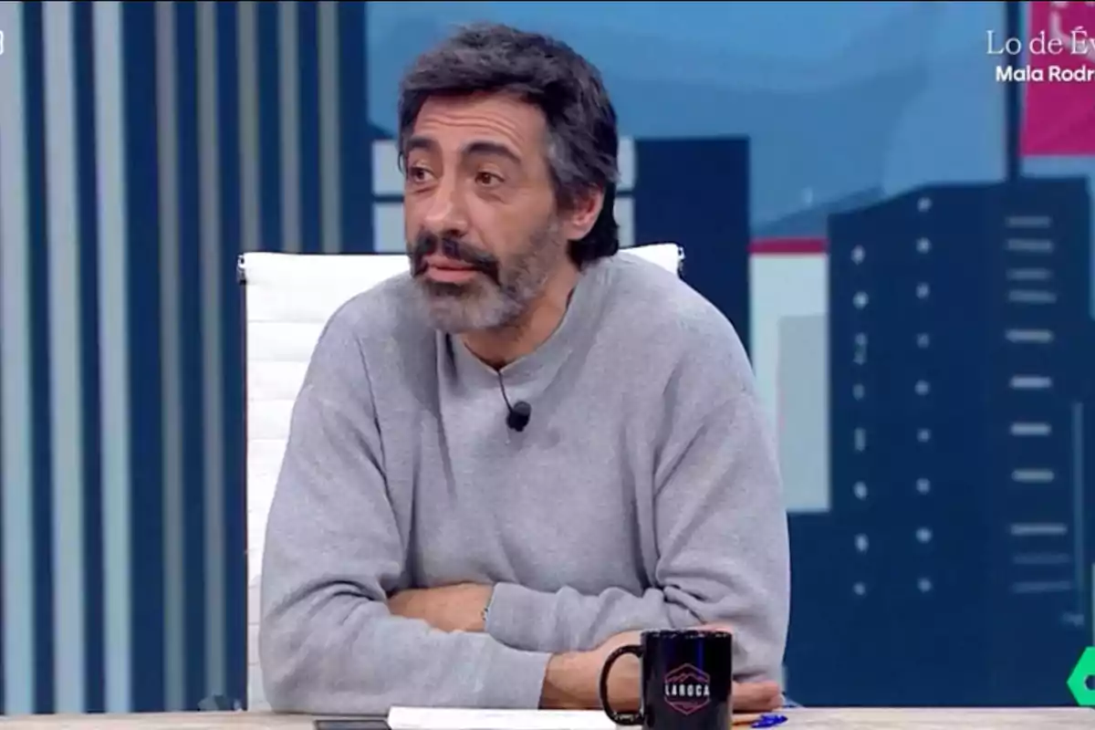 Un home amb barba i cabell fosc està assegut en un estudi de televisió, porta un jersei gris i té un micròfon de solapa, davant seu hi ha una tassa negra sobre la taula. Un home amb barba i cabell fosc està assegut en un estudi de televisió, porta un jersei gris i té un micròfon de solapa, davant seu hi ha una tassa negra sobre la taula.