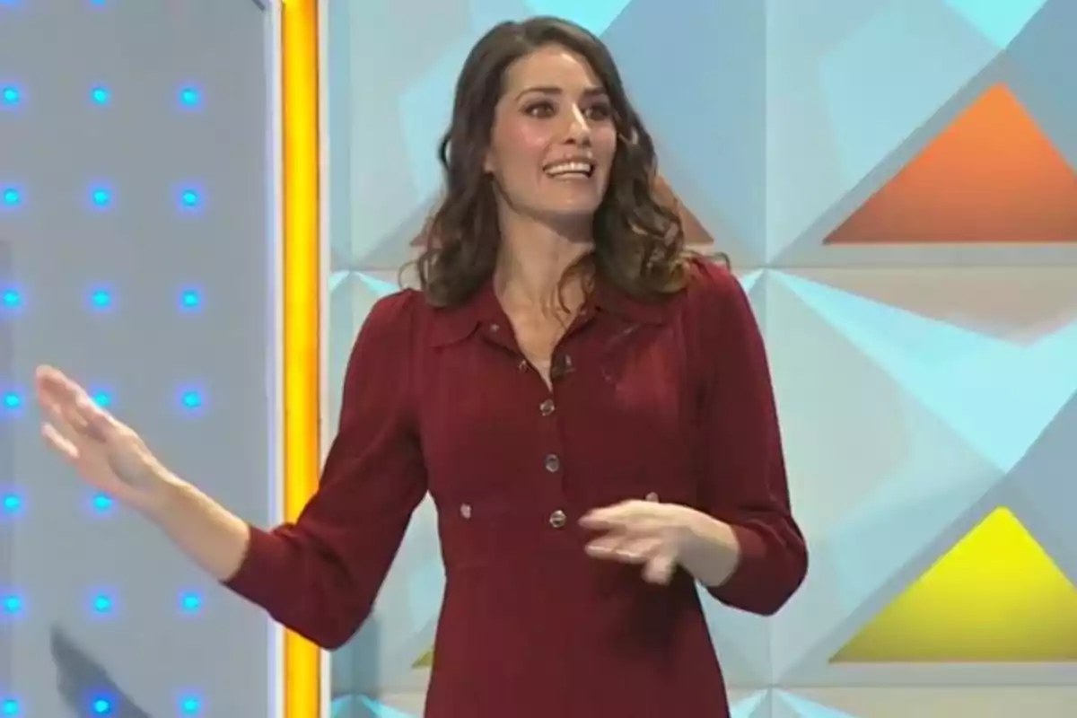 Laura Moure amb vestit vermell somriu mentre gesticula en un plató de televisió amb fons geomètric i llums blaves. Captura de 'La Ruleta de la Suerte' Laura Moure amb vestit vermell somriu mentre gesticula en un plató de televisió amb fons geomètric i llums blaves. Captura de 'La Ruleta de la Suerte'