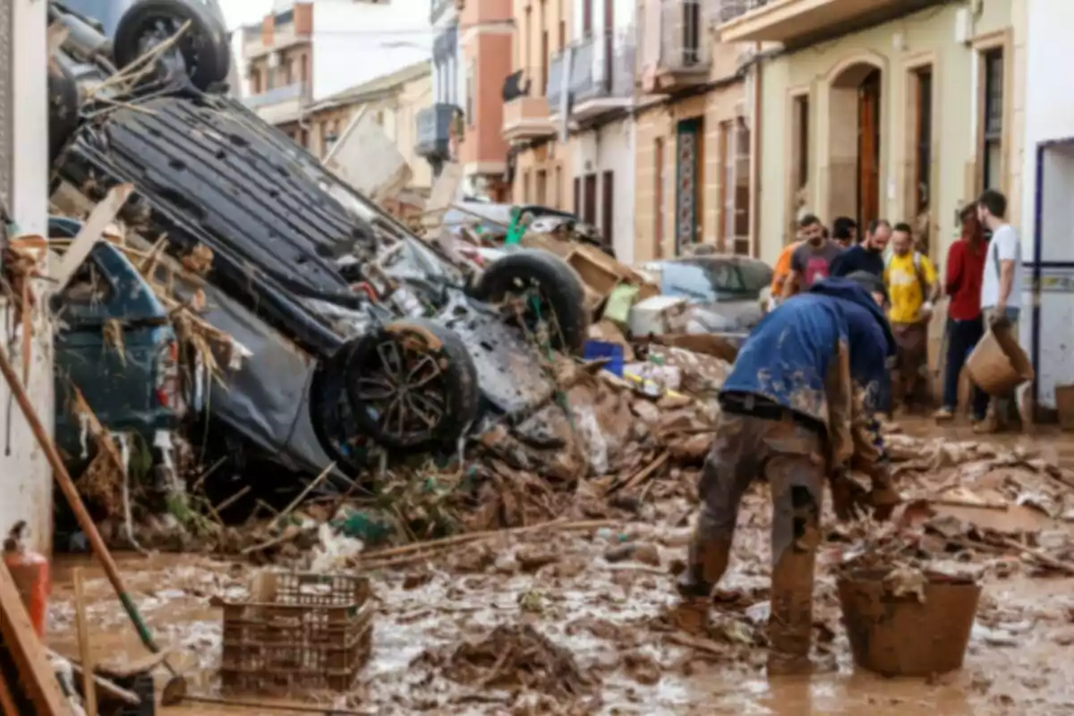 Un carrer ple de runes i fang després d'una inundació amb un cotxe bolcat i persones netejant.