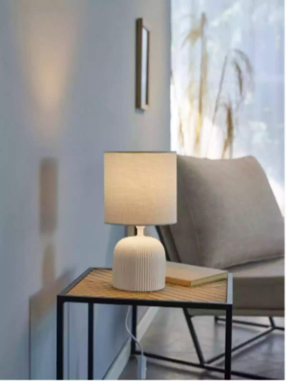 Làmpada de taula encesa sobre una taula auxiliar al costat d’un butacó en una sala il·luminada per llum natural Làmpada de taula encesa sobre una taula auxiliar al costat d’un butacó en una sala il·luminada per llum natural