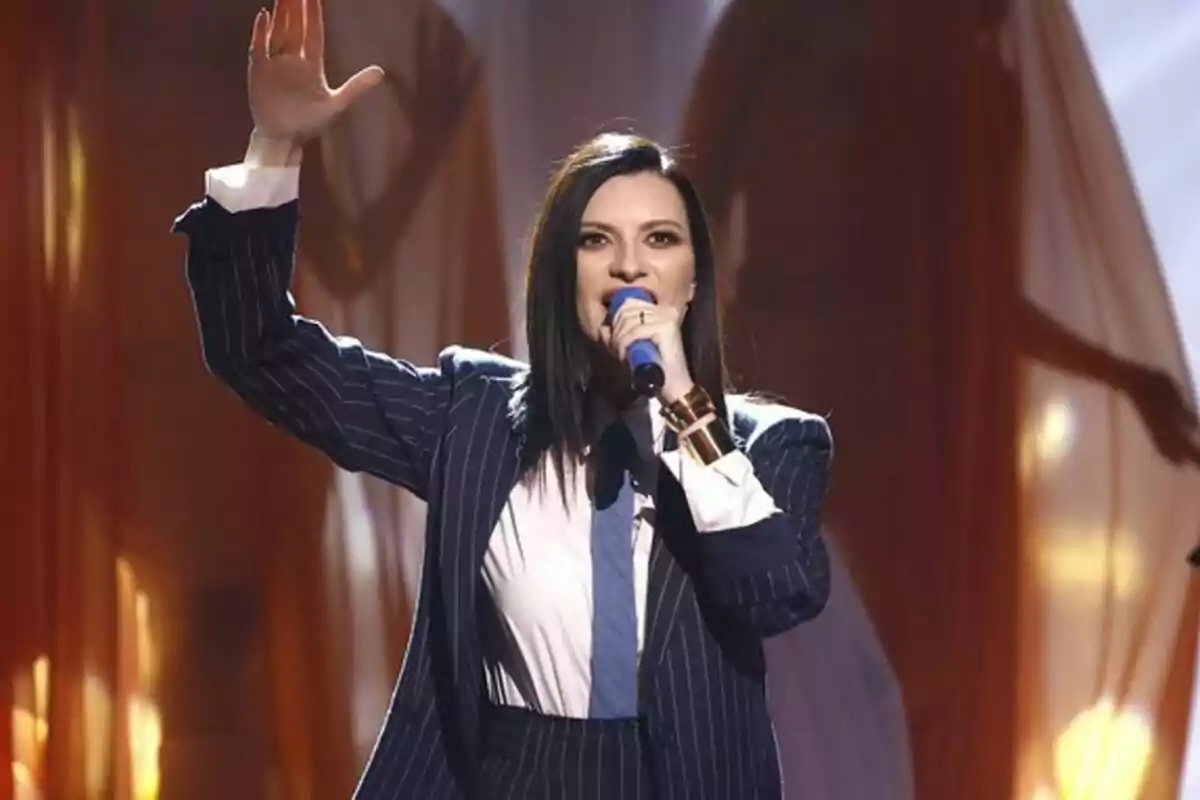 Laura Pausini con traje a rayas canta en un escenario iluminado mientras levanta una mano en La Voz. Laura Pausini con traje a rayas canta en un escenario iluminado mientras levanta una mano en La Voz.