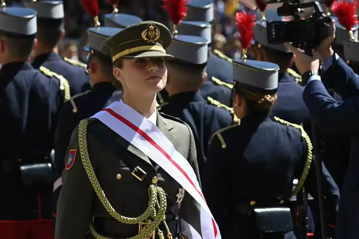 Leonor amb uniforme de gala i una banda blanca amb una franja vermella, envoltada d'altres militars en formació. Leonor amb uniforme de gala i una banda blanca amb una franja vermella, envoltada d'altres militars en formació.