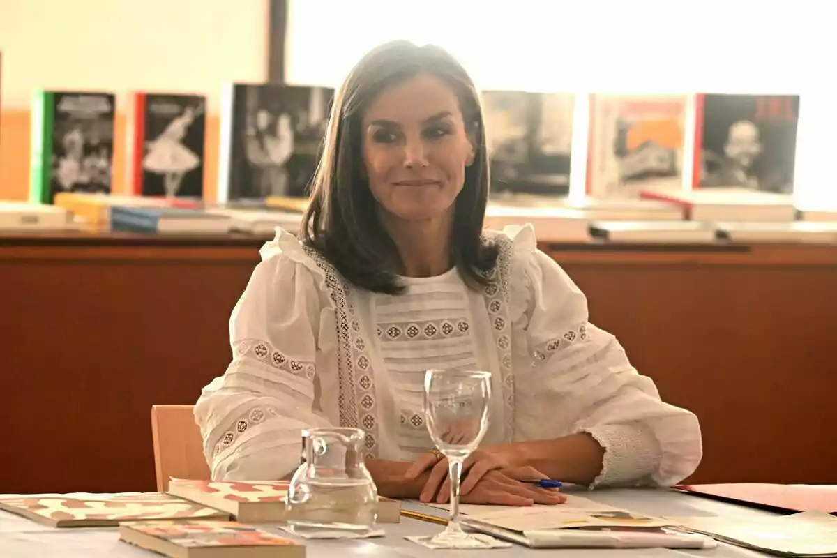 Letizia somrient mirant de banda amb una brusa blanca i les mans damunt la taula amb un bolígraf a la mà