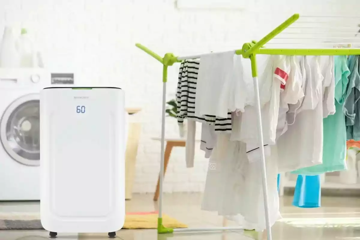Un deshumidificador blanc al costat d’un estenedor amb roba assecant-se en una habitació amb rentadora al fons Un deshumidificador blanc al costat d’un estenedor amb roba assecant-se en una habitació amb rentadora al fons