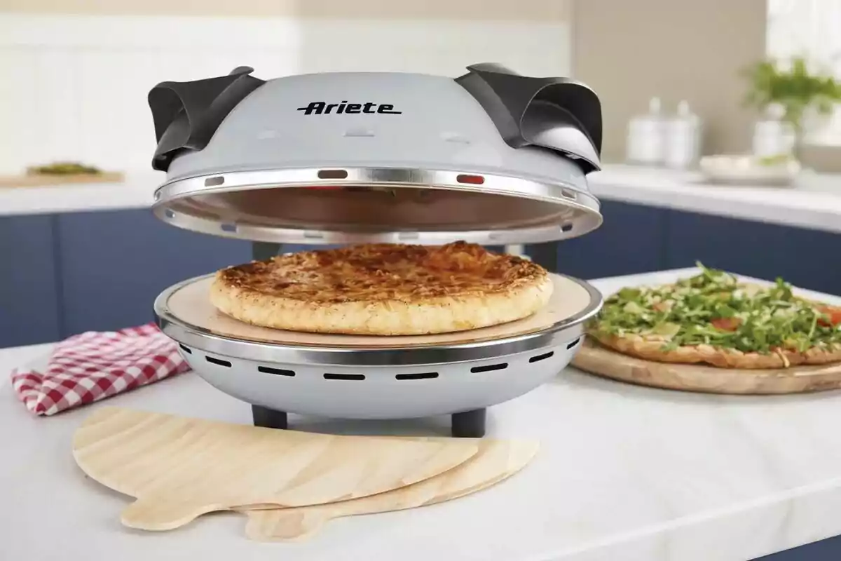 Una pizza cuinant-se en un forn elèctric sobre un taulell de cuina, amb una pizza addicional coberta de rúcula al fons i un drap de cuina de quadres vermells i blancs al costat. Una pizza cuinant-se en un forn elèctric sobre un taulell de cuina, amb una pizza addicional coberta de rúcula al fons i un drap de cuina de quadres vermells i blancs al costat.