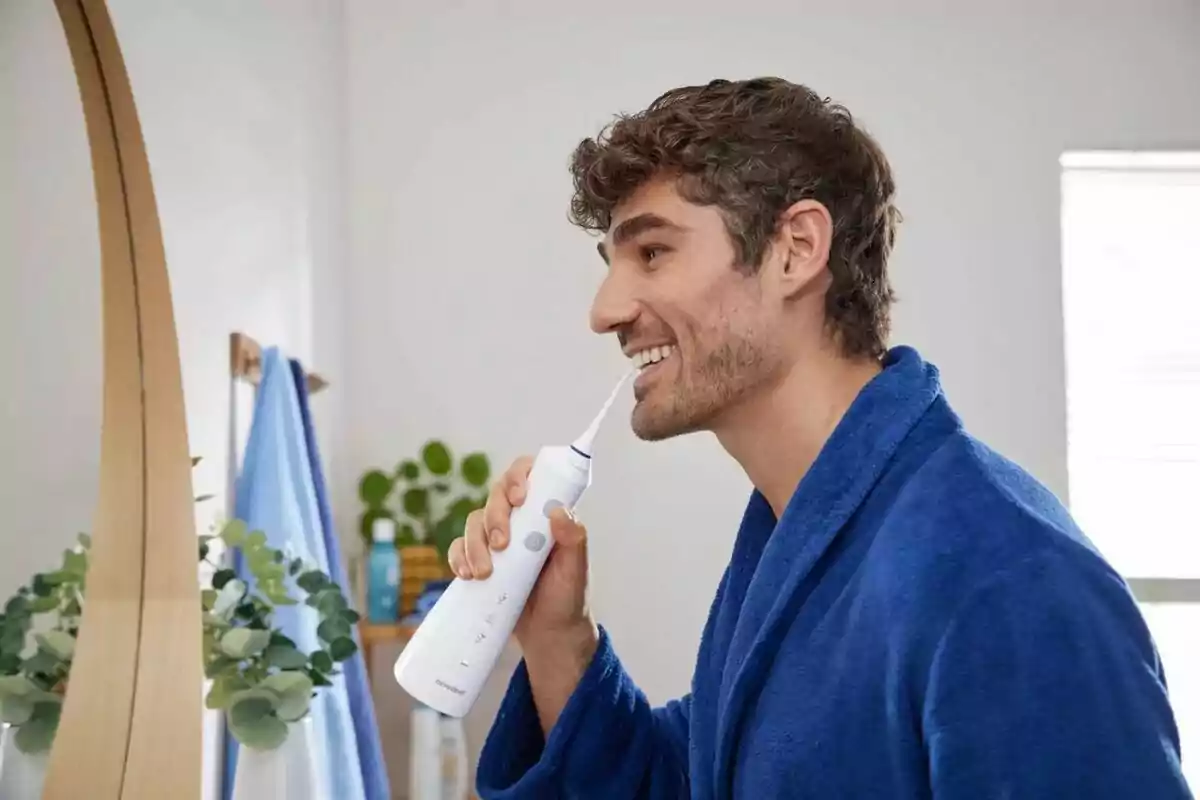 Home usant un irrigador dental davant del mirall al bany, vestint una bata blava. Home usant un irrigador dental davant del mirall al bany, vestint una bata blava.