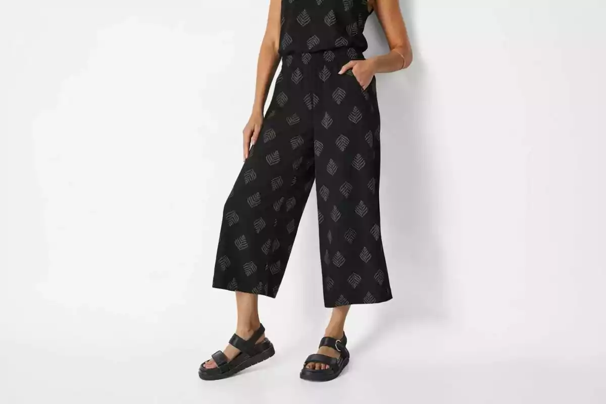 Dona amb un mono negre amb estampat de fulles i sandàlies negres sobre fons blanc Dona amb un mono negre amb estampat de fulles i sandàlies negres sobre fons blanc