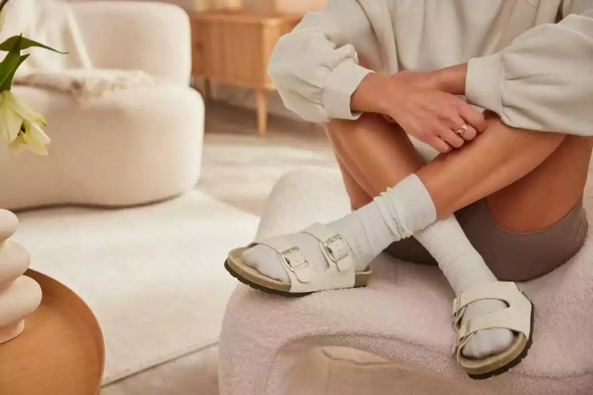 Persona asseguda en una cadira moderna amb sandàlies blanques i mitjons, en una sala d’estar de tons clars i decoració minimalista. Persona asseguda en una cadira moderna amb sandàlies blanques i mitjons, en una sala d’estar de tons clars i decoració minimalista.