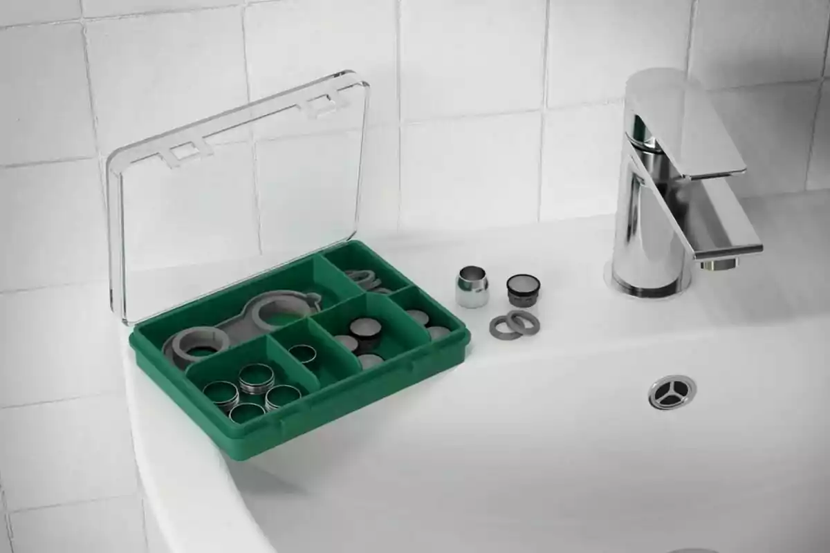 Caixa verda amb compartiments que conté peces i eines petites sobre un lavabo blanc al costat d’una aixeta de bany platejada. Caixa verda amb compartiments que conté peces i eines petites sobre un lavabo blanc al costat d’una aixeta de bany platejada.