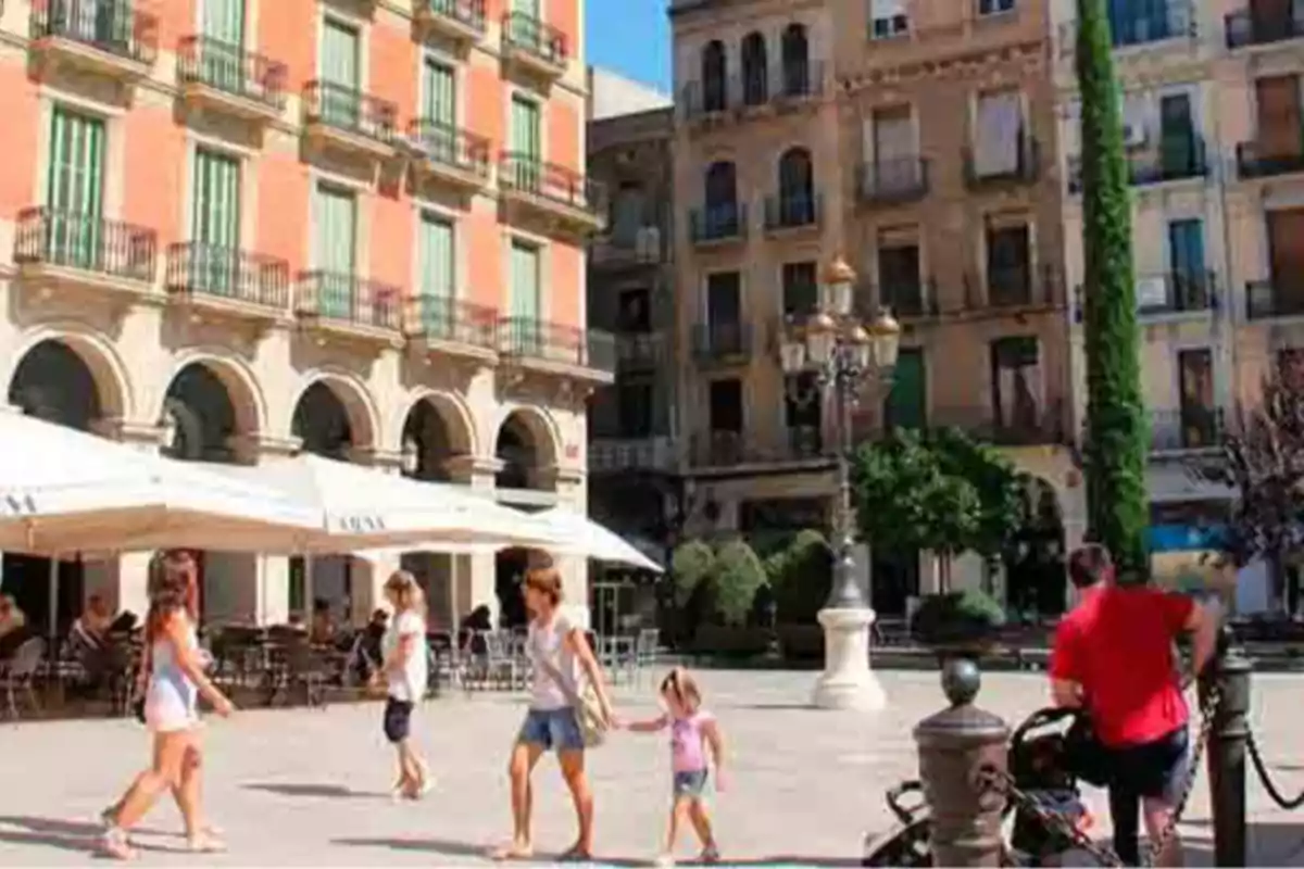 Una plaça assolellada amb edificis antics i persones caminant a prop d'una terrassa amb para-sols blancs.