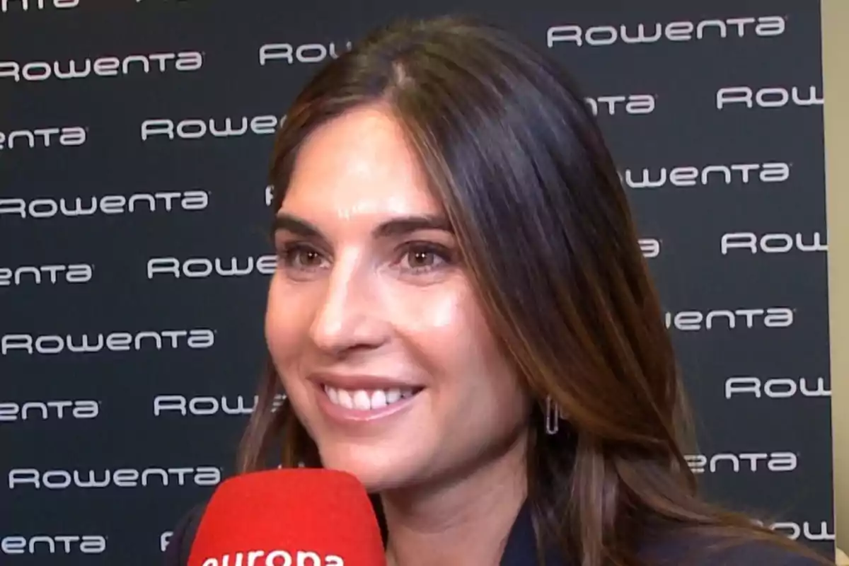 Lourdes Montes somrient sosté un micròfon vermell davant d'un fons amb el logotip de Rowenta repetit. Lourdes Montes somrient sosté un micròfon vermell davant d'un fons amb el logotip de Rowenta repetit.
