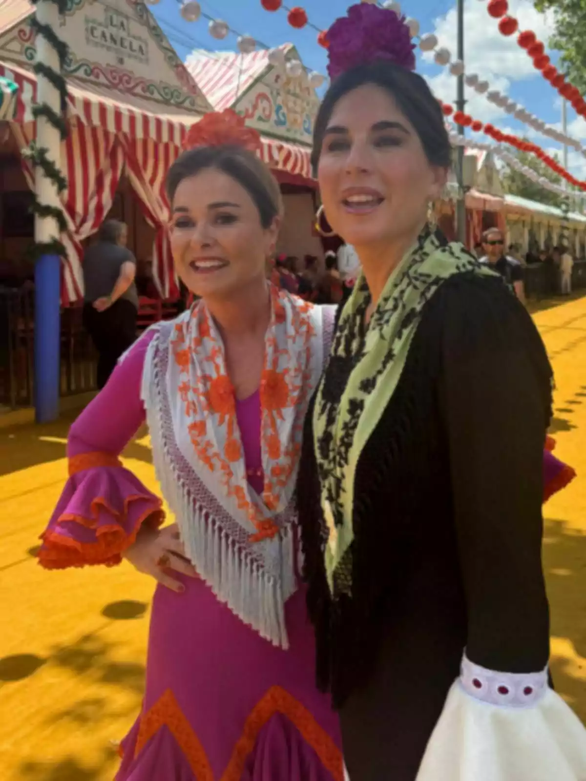 Lourdes Montes i una altra dona vestides amb vestits tradicionals posen somrients en un esdeveniment festiu a l'aire lliure, amb casetes decorades i fanalets de colors al fons. Lourdes Montes i una altra dona vestides amb vestits tradicionals posen somrients en un esdeveniment festiu a l'aire lliure, amb casetes decorades i fanalets de colors al fons.
