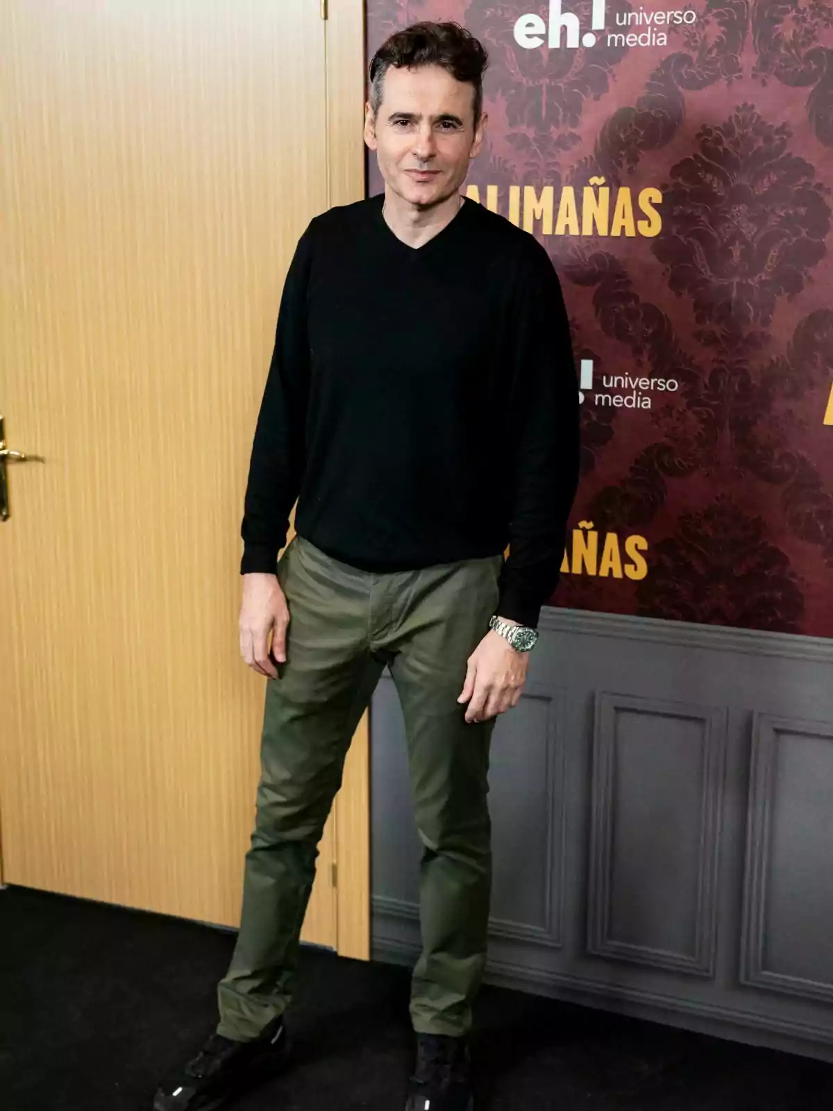 Luis Miguel Segui amb suèter negre i pantalons verds posant davant d'un fons decoratiu.