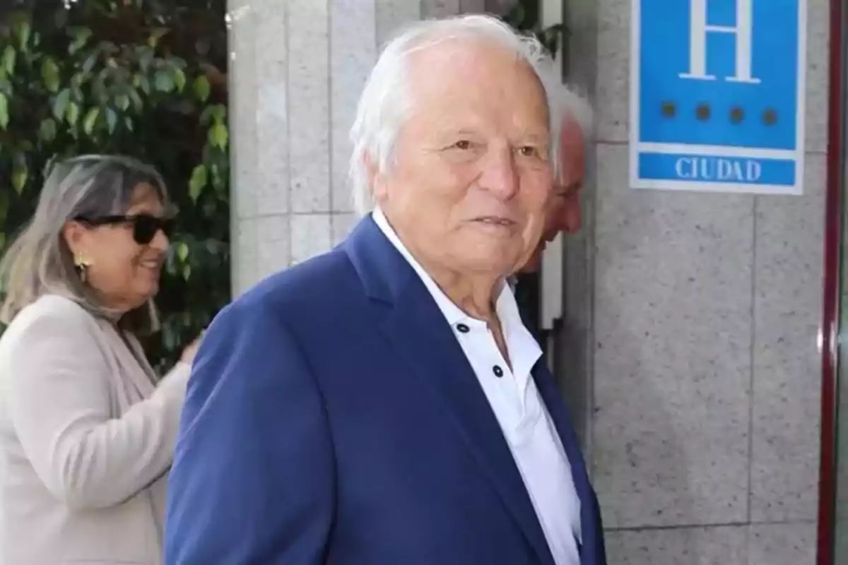 Manuel Benítez amb cabell blanc i vestit blau està dret davant d'un edifici amb una dona somrient al fons. Manuel Benítez amb cabell blanc i vestit blau està dret davant d'un edifici amb una dona somrient al fons.