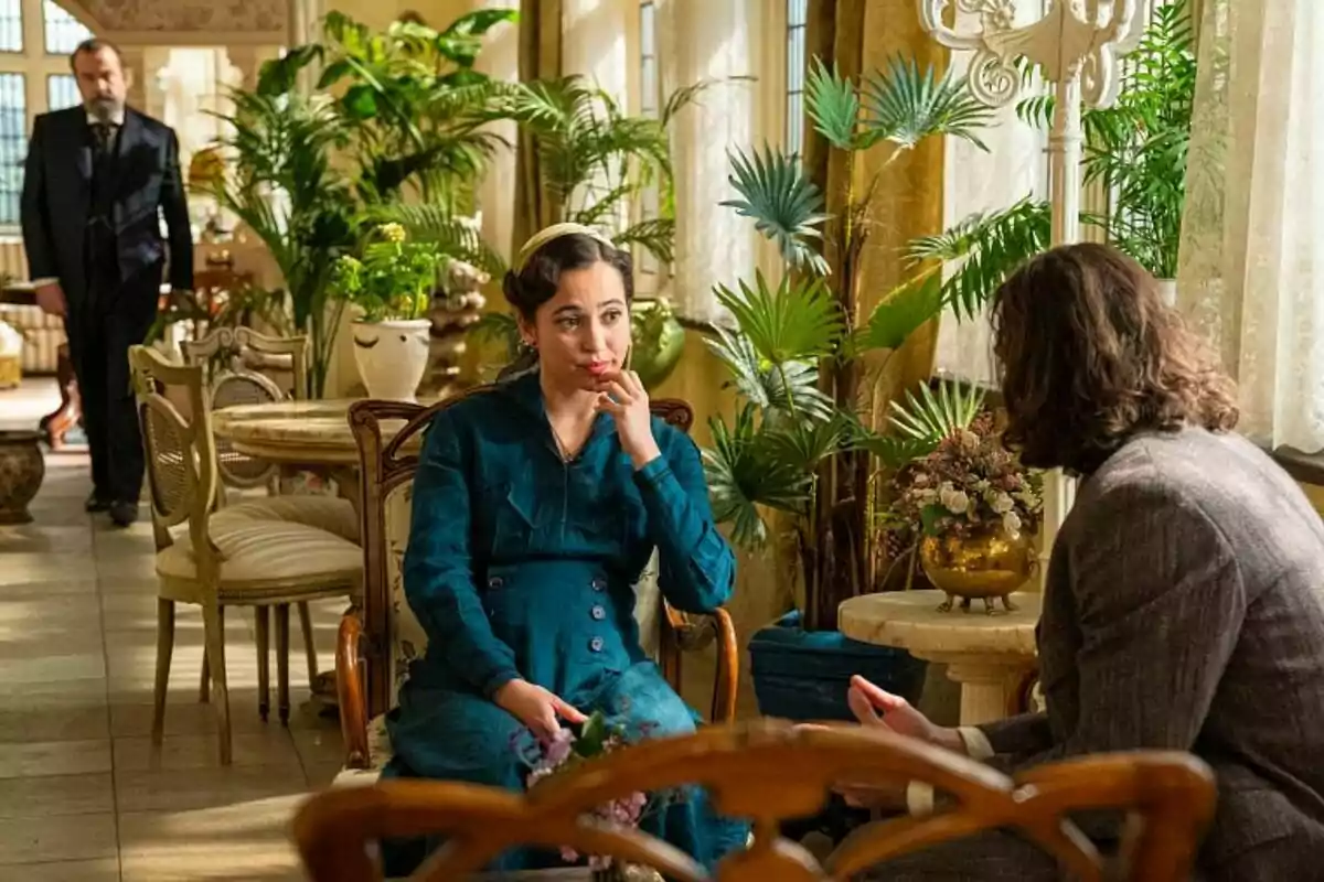 Carmen Asecas amb vestit blau està asseguda en una cadira en una habitació decorada amb plantes mentre conversa amb Ibrahim Al Shami de cabell llarg; al fons, Manuel Regueiro observa l'escena a La Promesa. Carmen Asecas amb vestit blau està asseguda en una cadira en una habitació decorada amb plantes mentre conversa amb Ibrahim Al Shami de cabell llarg; al fons, Manuel Regueiro observa l'escena a La Promesa.