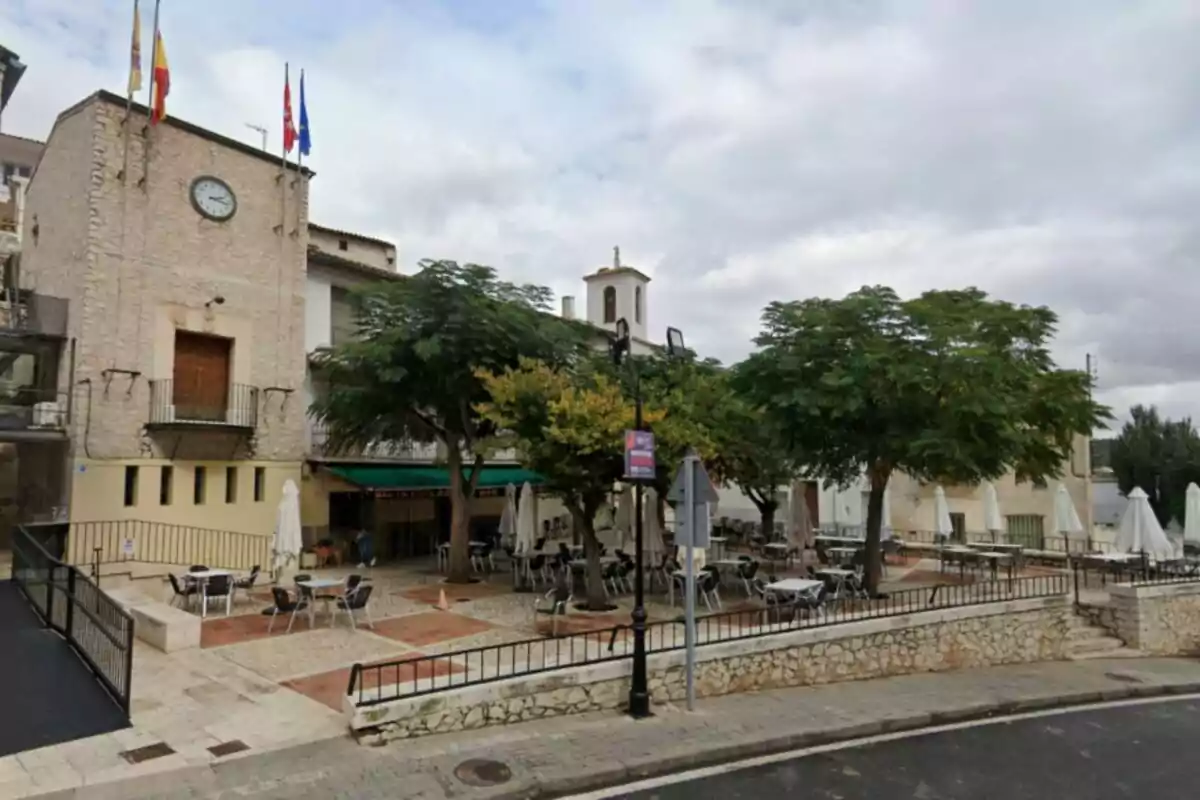 Una plaça amb taules i cadires a l'aire lliure envoltada d'arbres i un edifici de pedra amb un rellotge i diverses banderes a la part superior. Una plaça amb taules i cadires a l'aire lliure envoltada d'arbres i un edifici de pedra amb un rellotge i diverses banderes a la part superior.