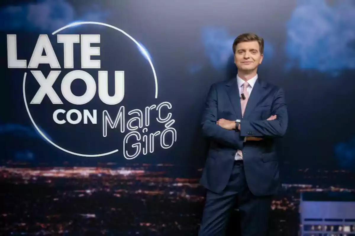 Marc Giró amb un vestit posa amb els braços plegats davant d'un fons que publicita el seu programa, "Late Xou amb Marc Giró".