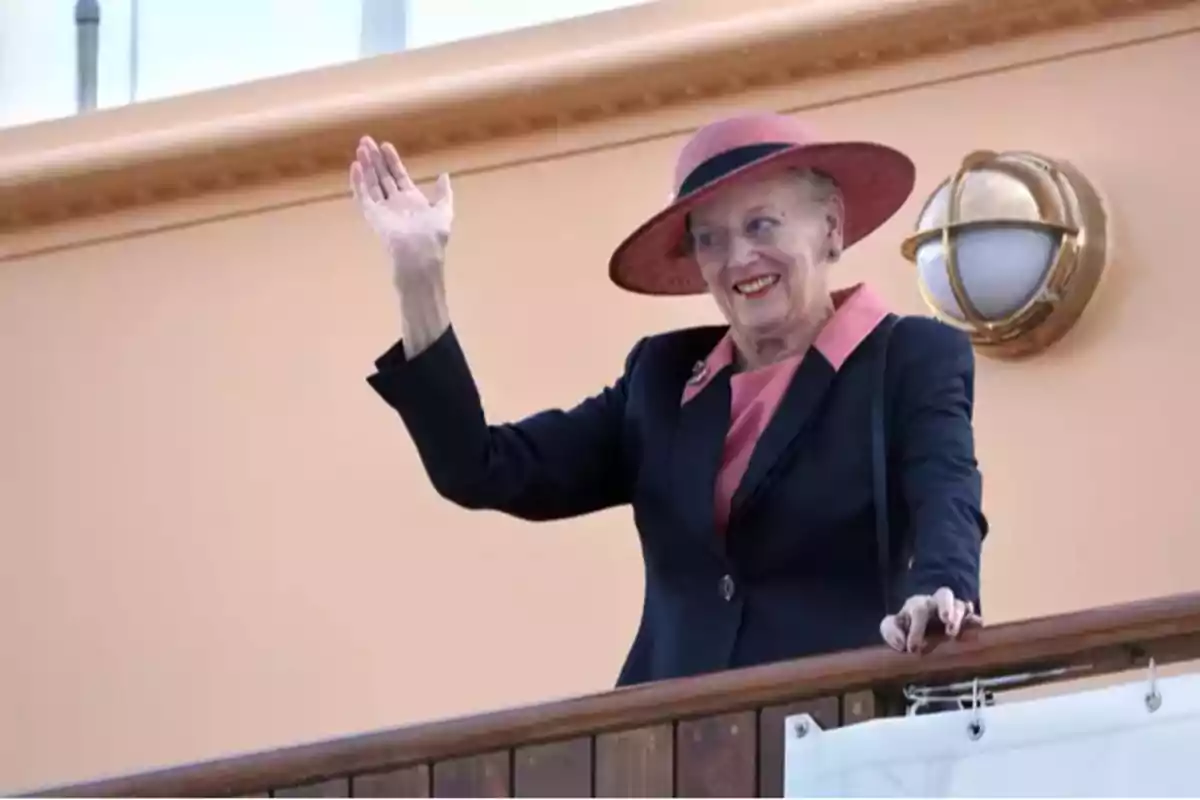 Queen Margaret of Denmark in a pink hat and dark suit waves from a balcony. Queen Margaret of Denmark in a pink hat and dark suit waves from a balcony.