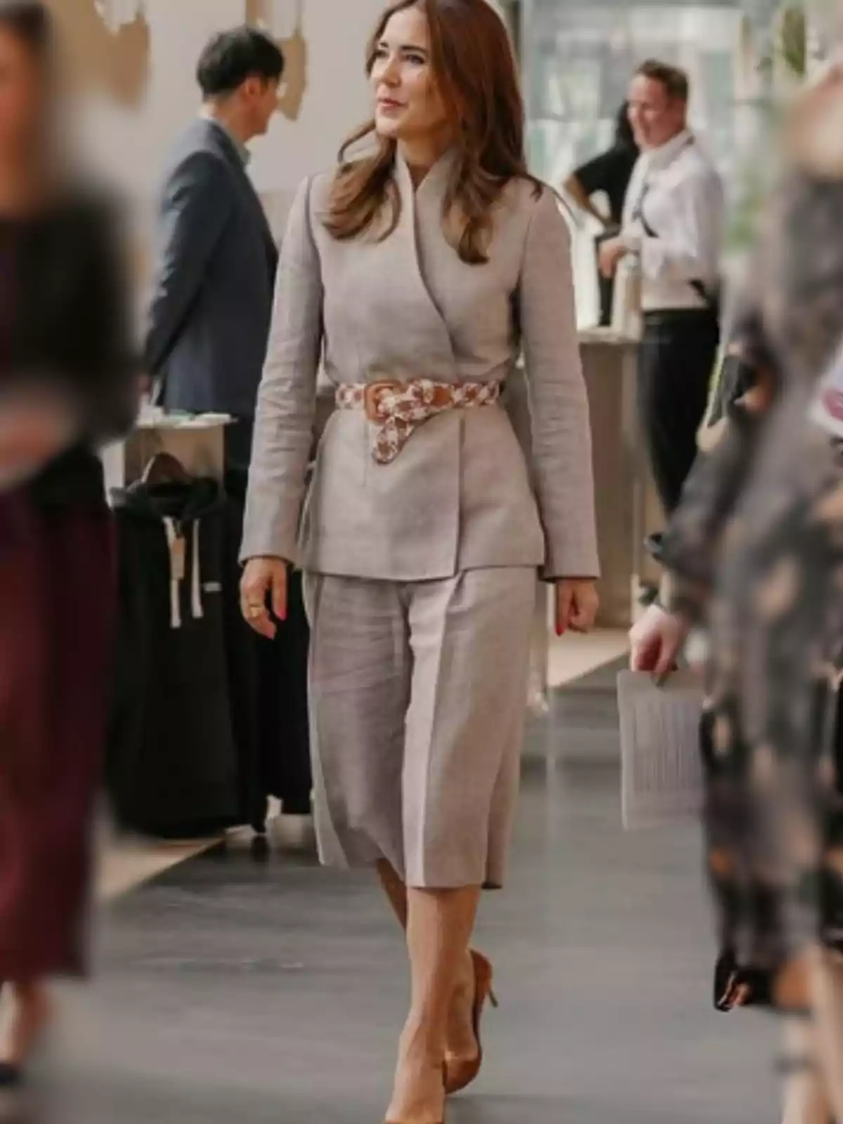 Dona amb cabell castany clar portant un conjunt beix de jaqueta i faldilla midi, cinturó estampat i talons nude, caminant en un espai interior amb altres persones al voltant. Dona amb cabell castany clar portant un conjunt beix de jaqueta i faldilla midi, cinturó estampat i talons nude, caminant en un espai interior amb altres persones al voltant.