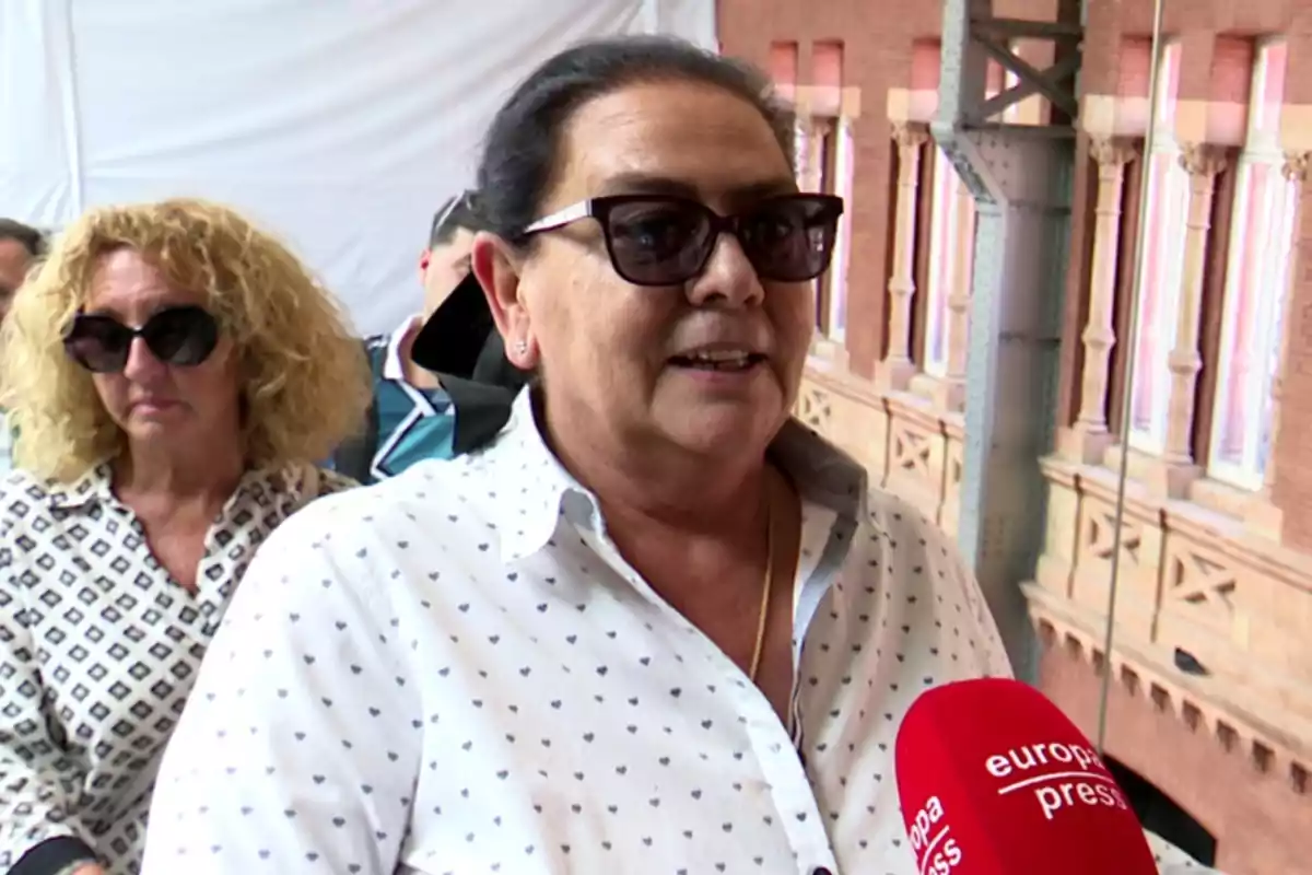 María del Monte amb ulleres de sol i camisa blanca amb petits estampats sent entrevistada per un micròfon vermell d’Europa Press, amb una altra dona al fons i un edifici de maó darrere. María del Monte amb ulleres de sol i camisa blanca amb petits estampats sent entrevistada per un micròfon vermell d’Europa Press, amb una altra dona al fons i un edifici de maó darrere.