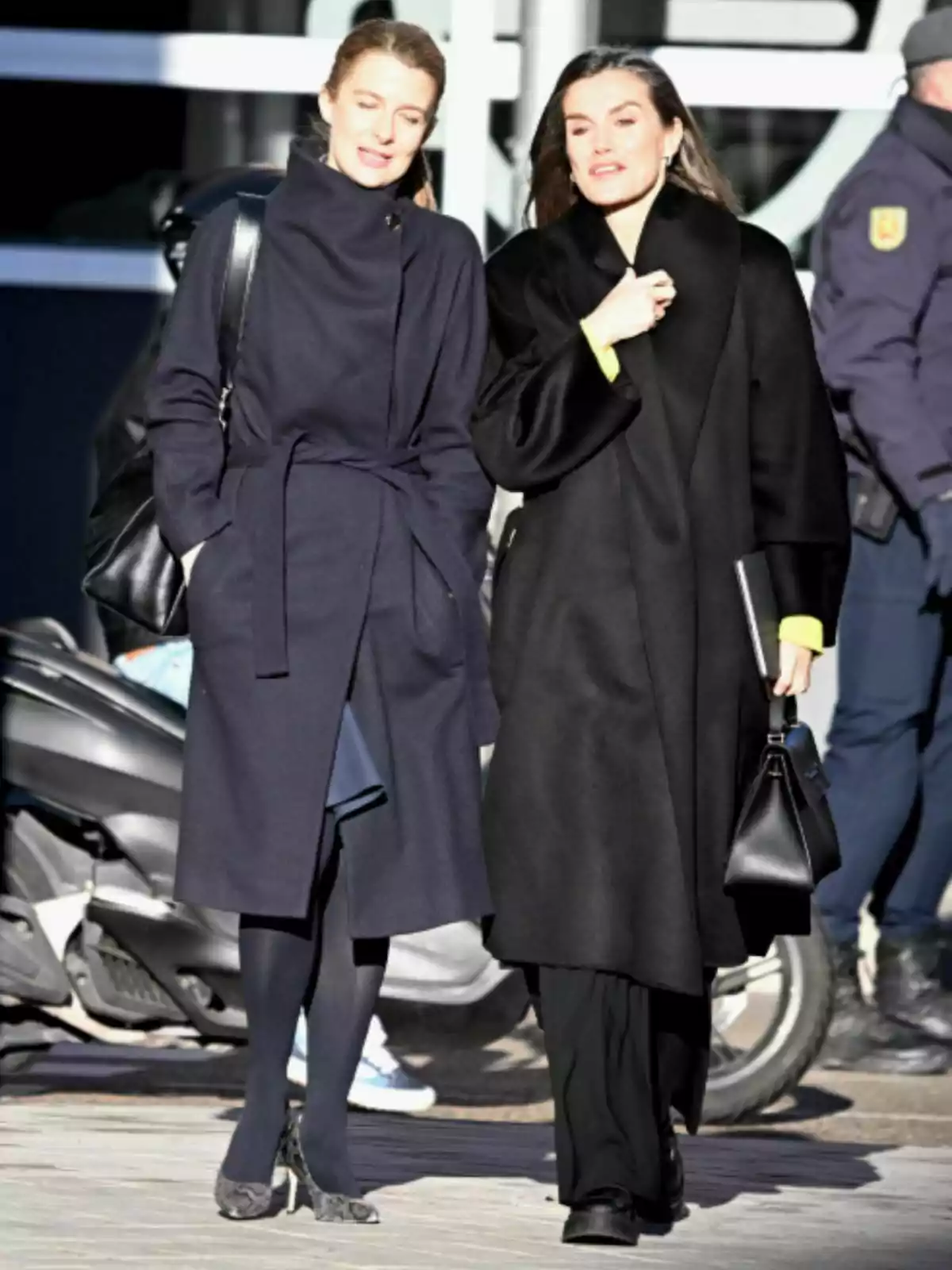 María Dolores Ocaña and Letizia walking together, dressed in dark coats, one of them carrying a black purse and a notebook. María Dolores Ocaña and Letizia walking together, dressed in dark coats, one of them carrying a black purse and a notebook.