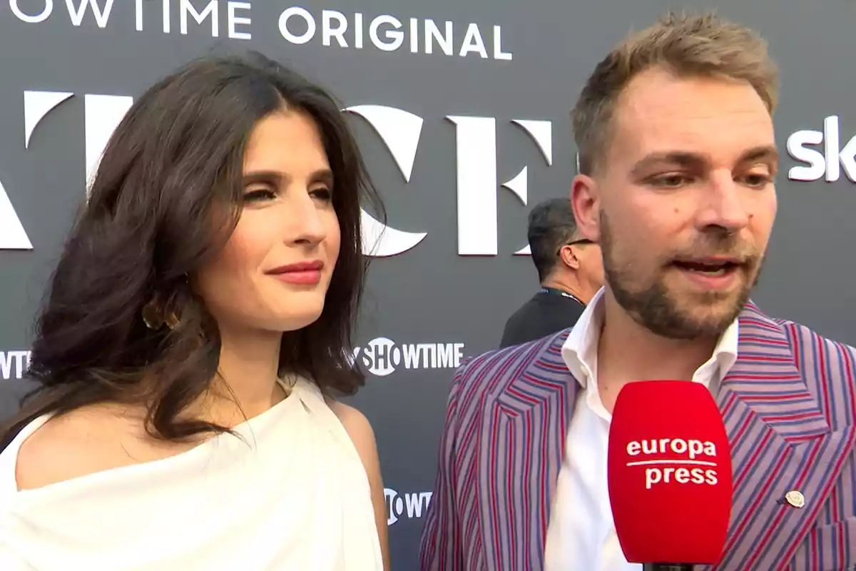 María Sánchez amb vestit blanc i José María Almoguera amb jaqueta de ratlles parlen davant d’un micròfon vermell d’Europa Press en un esdeveniment amb un fons de lletres grans. María Sánchez amb vestit blanc i José María Almoguera amb jaqueta de ratlles parlen davant d’un micròfon vermell d’Europa Press en un esdeveniment amb un fons de lletres grans.