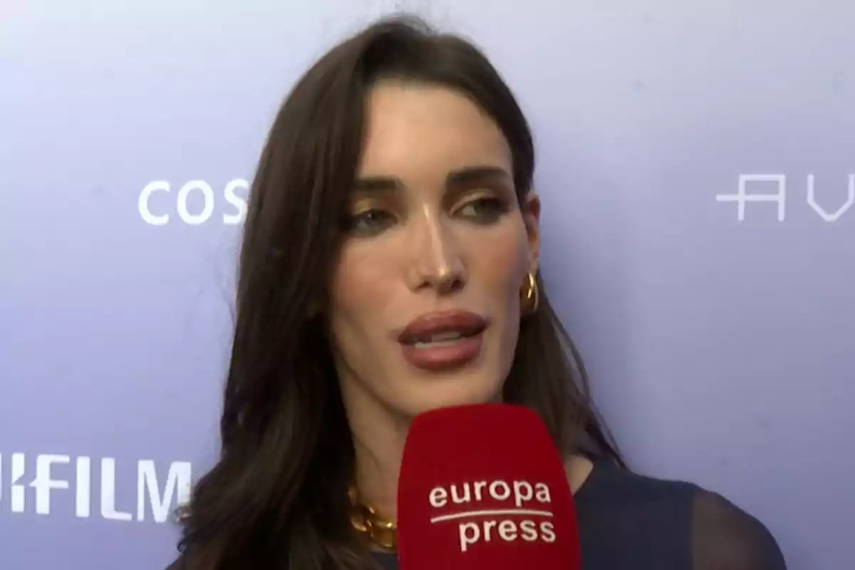 Marta López Álamo donant una entrevista amb un micròfon vermell d’Europa Press davant d’un fons lila amb logotips blancs