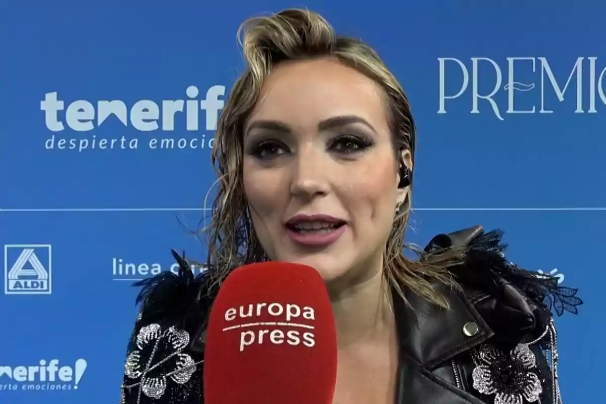 Marta Riesco amb jaqueta negra decorada sosté un micròfon vermell d'Europa Press davant d'un fons blau amb logotips de Tenerife i Aldi. Marta Riesco amb jaqueta negra decorada sosté un micròfon vermell d'Europa Press davant d'un fons blau amb logotips de Tenerife i Aldi.