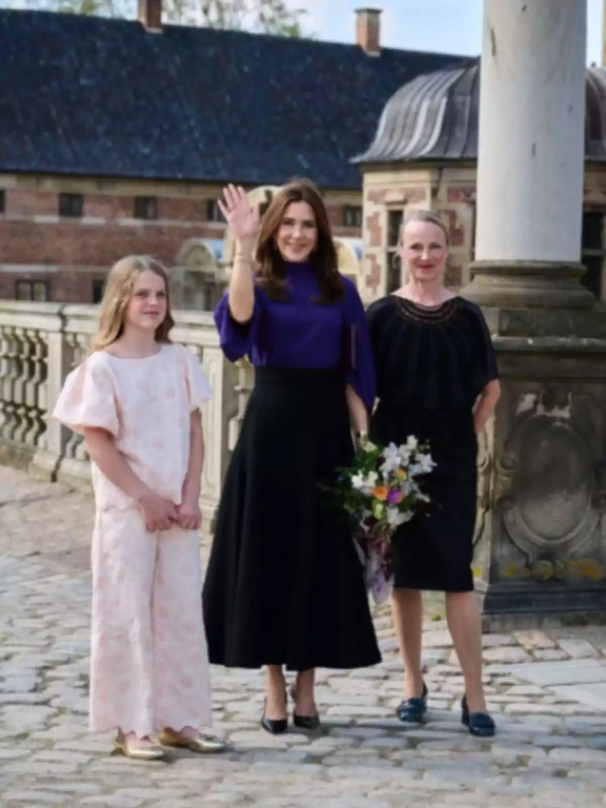 Tres persones posen a l'aire lliure en un entorn històric, una d'elles saluda mentre sosté un ram de flors. Tres persones posen a l'aire lliure en un entorn històric, una d'elles saluda mentre sosté un ram de flors.