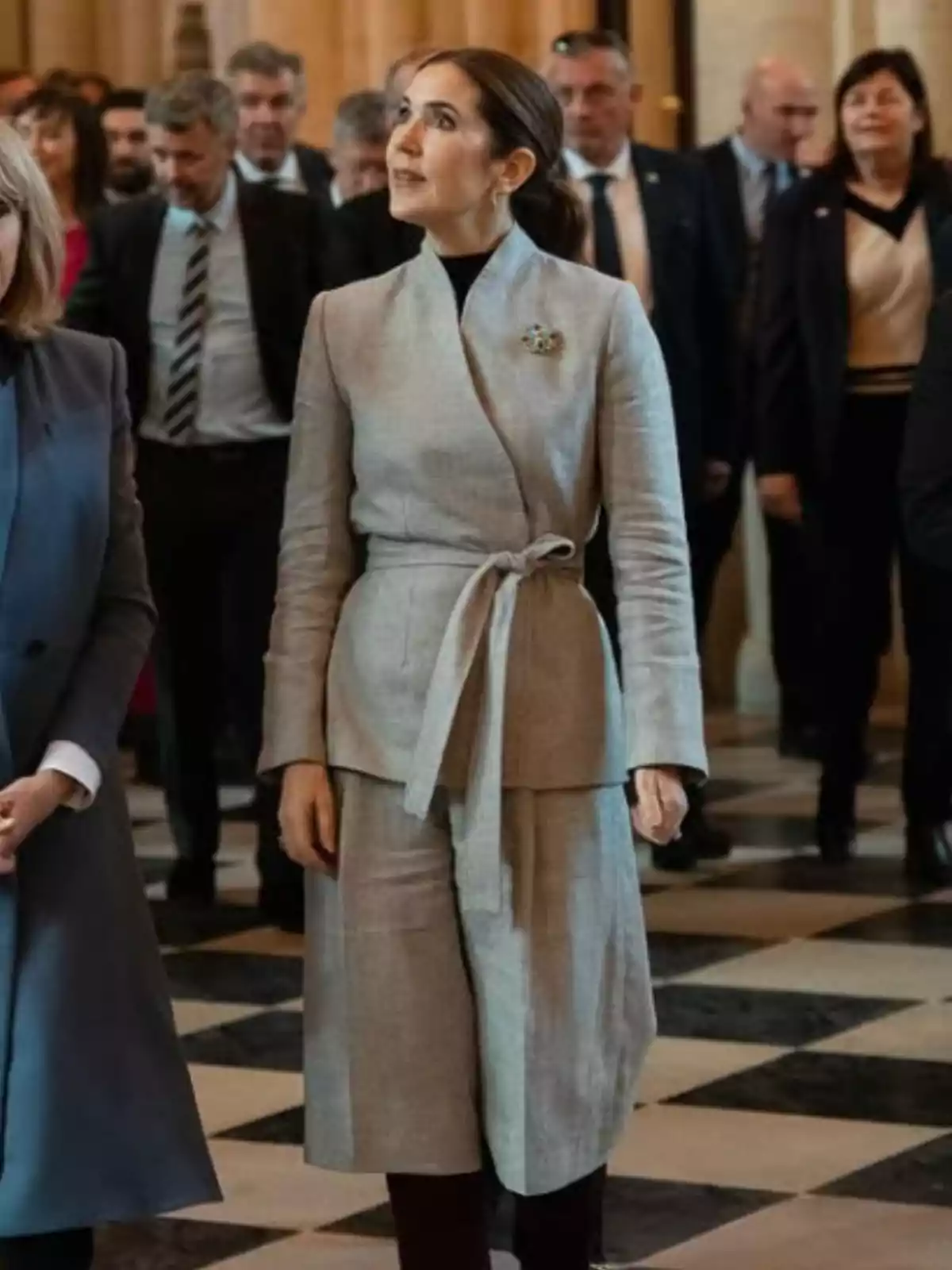 Dona vestida amb un conjunt gris clar i un fermall, dreta en un lloc elegant amb terra de rajoles quadrades, envoltada de diverses persones amb vestit formal. Dona vestida amb un conjunt gris clar i un fermall, dreta en un lloc elegant amb terra de rajoles quadrades, envoltada de diverses persones amb vestit formal.