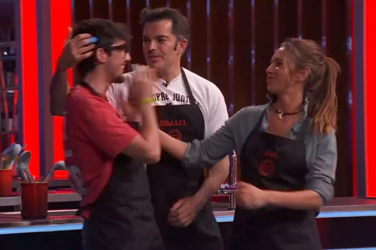 Emilio, Ismael i Ana amb davantals de cuina interactuen en un plató de competició culinària, una dona somriu mentre toca la cara d’un home amb ulleres i un altre home els observa. Captura de 'MasterChef' Emilio, Ismael i Ana amb davantals de cuina interactuen en un plató de competició culinària, una dona somriu mentre toca la cara d’un home amb ulleres i un altre home els observa. Captura de 'MasterChef'
