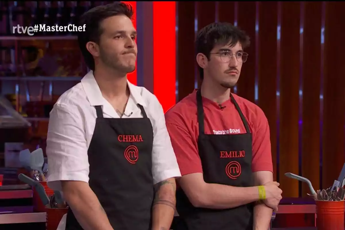Dos concursants de MasterChef amb davantals negres i noms brodats en vermell estan drets a la cuina del programa amb una expressió seriosa. Dos concursants de MasterChef amb davantals negres i noms brodats en vermell estan drets a la cuina del programa amb una expressió seriosa.
