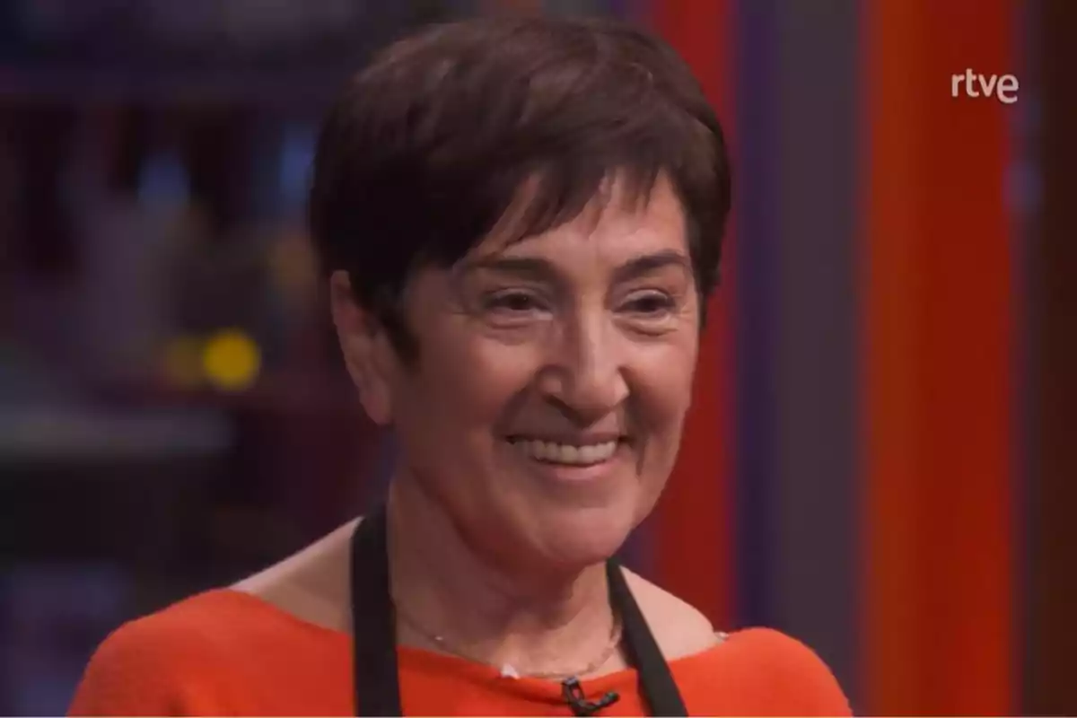 Ana María somrient amb cabell curt i fosc porta un micròfon de solapa i un davantal negre sobre una peça vermella, amb un fons desenfocat i el logotip de RTVE a la cantonada superior dreta. Captura de 'MasterChef' Ana María somrient amb cabell curt i fosc porta un micròfon de solapa i un davantal negre sobre una peça vermella, amb un fons desenfocat i el logotip de RTVE a la cantonada superior dreta. Captura de 'MasterChef'