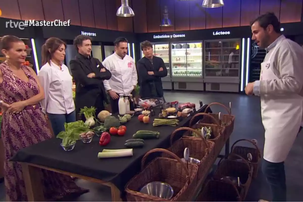 Un grup de persones en un plató de cuina d’un programa de televisió observen una taula amb ingredients frescos i cistelles de vímet; al fons es veuen neveres amb productes lactis i embotits. Captura de ‘MasterChef’. Un grup de persones en un plató de cuina d’un programa de televisió observen una taula amb ingredients frescos i cistelles de vímet; al fons es veuen neveres amb productes lactis i embotits. Captura de ‘MasterChef’.