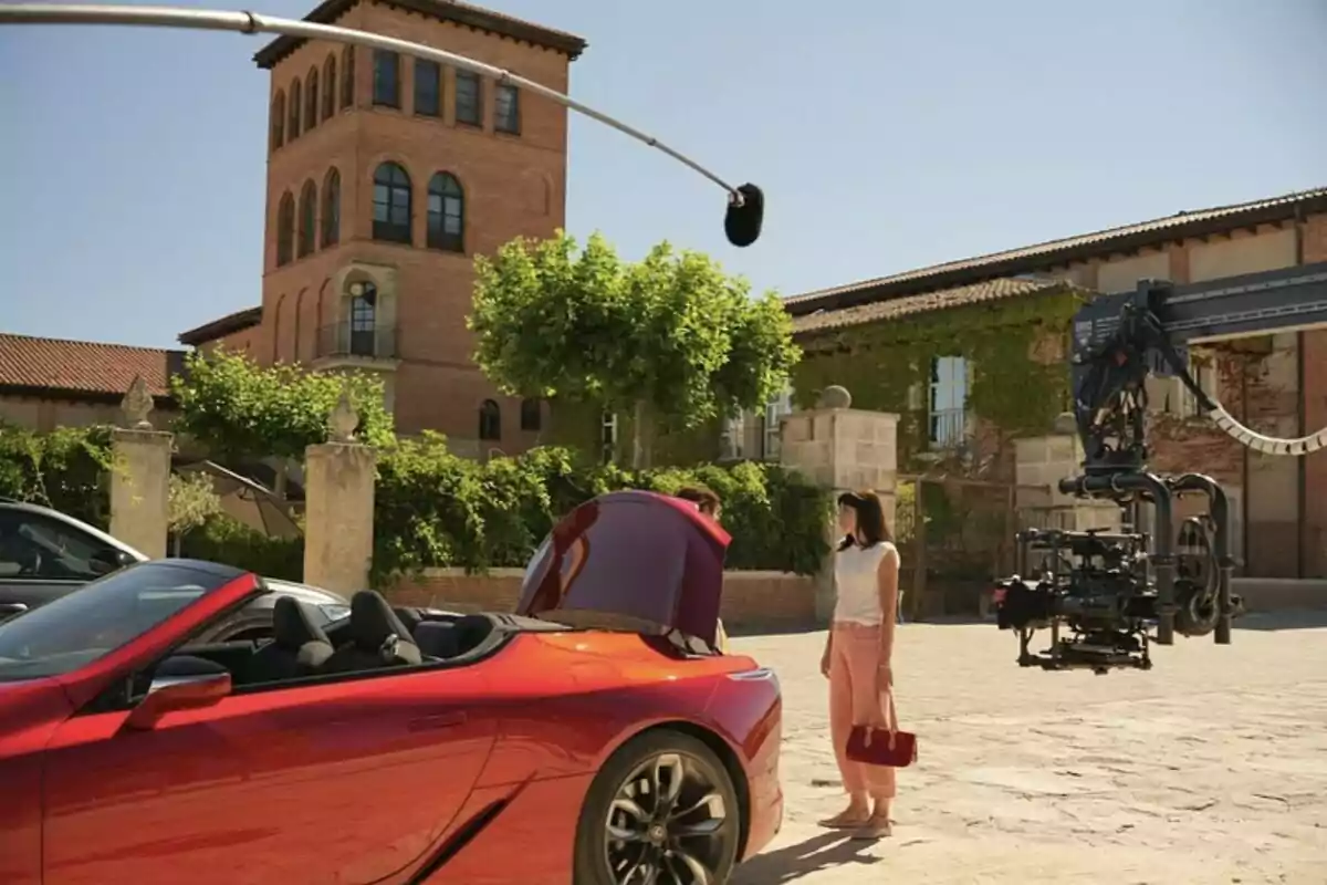 A woman standing next to a red sports car with the trunk open while a film crew with a camera and microphone records in an outdoor setting with a brick building in the background. A woman standing next to a red sports car with the trunk open while a film crew with a camera and microphone records in an outdoor setting with a brick building in the background.