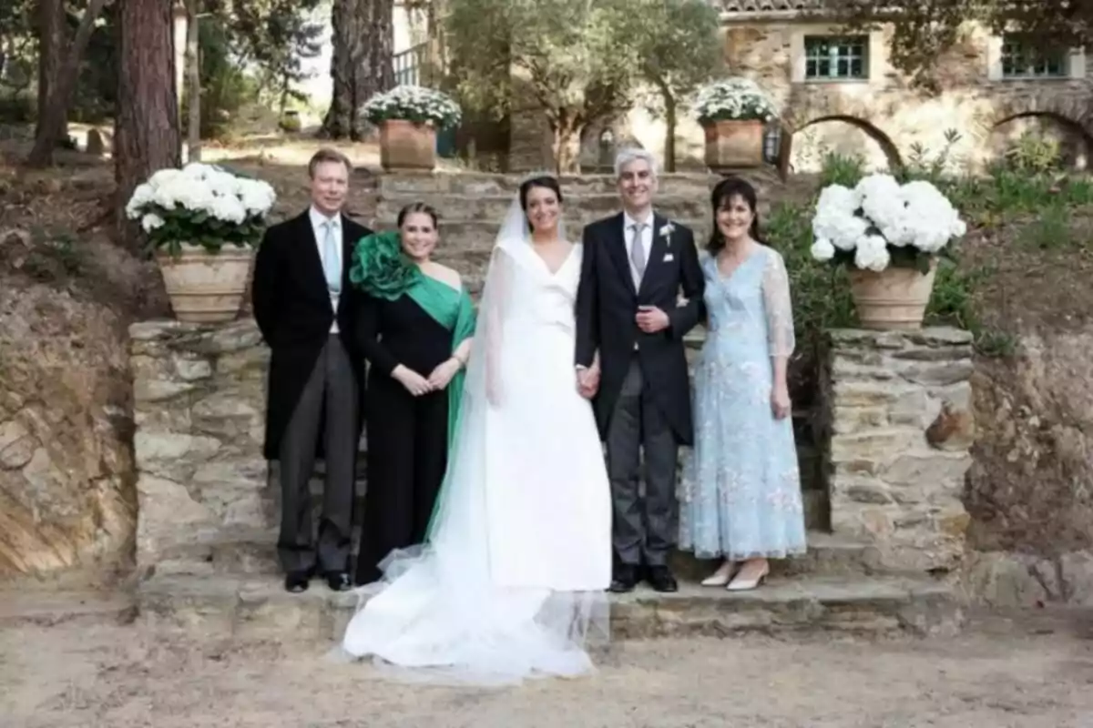 Un grup de persones elegantment vestides posant per a una foto en un entorn a l'aire lliure amb escales de pedra i testos amb flors blanques. Un grup de persones elegantment vestides posant per a una foto en un entorn a l'aire lliure amb escales de pedra i testos amb flors blanques.