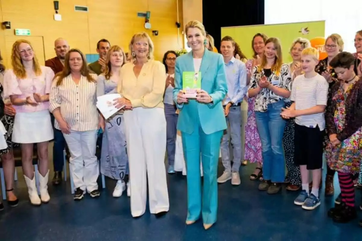 Un grup de persones dretes en una sala, algunes aplaudint, mentre dues dones al centre sostenen documents i somriuen. Un grup de persones dretes en una sala, algunes aplaudint, mentre dues dones al centre sostenen documents i somriuen.