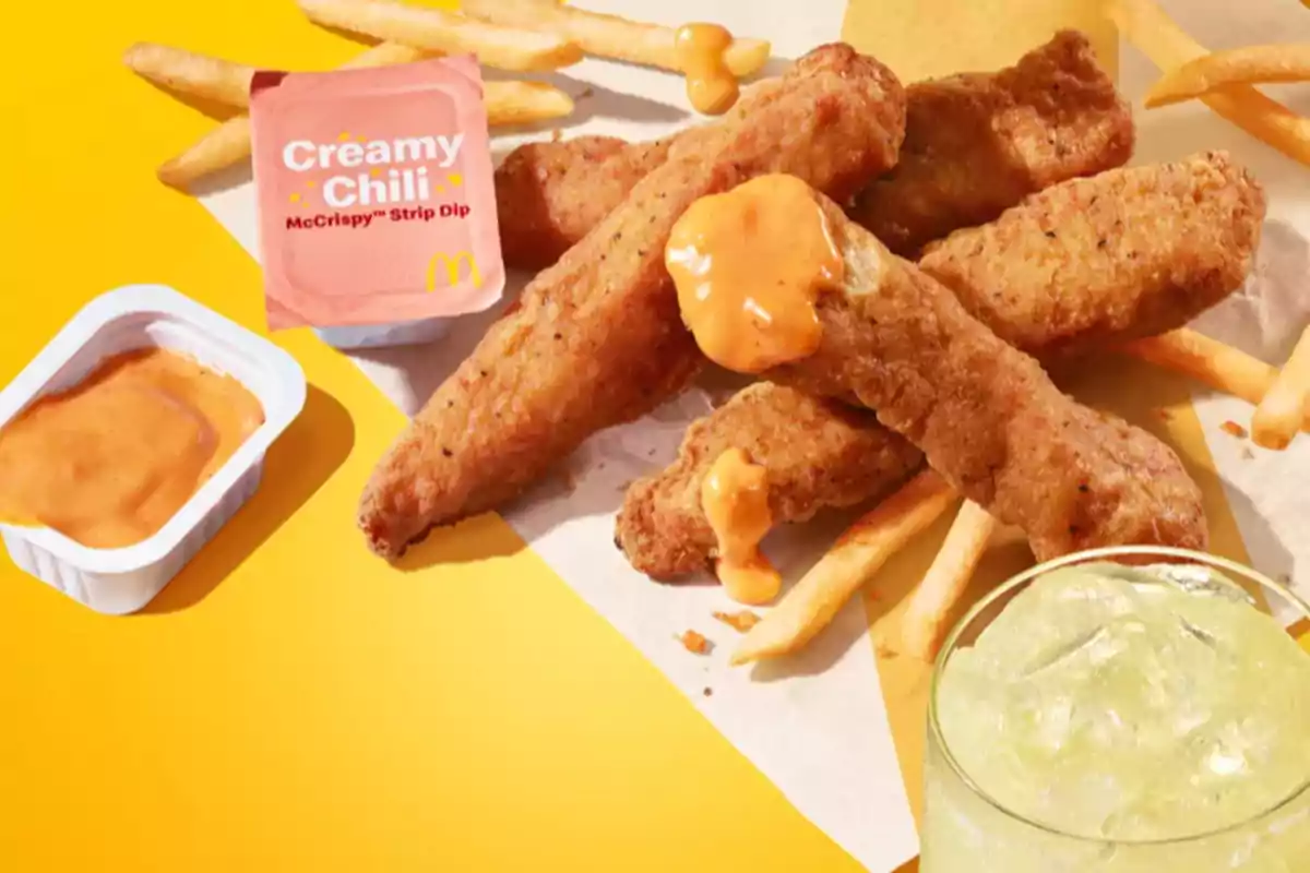 Plato de tiras de pollo empanizadas con papas fritas, acompañadas de una salsa cremosa de chile y una bebida con hielo sobre un fondo amarillo. Plato de tiras de pollo empanizadas con papas fritas, acompañadas de una salsa cremosa de chile y una bebida con hielo sobre un fondo amarillo.