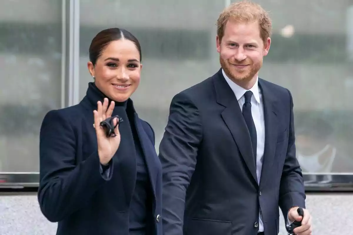 Meghan Markle i el príncep Harry vestits de manera formal, tots dos somrient i caminant junts, ella està saludant amb la mà.