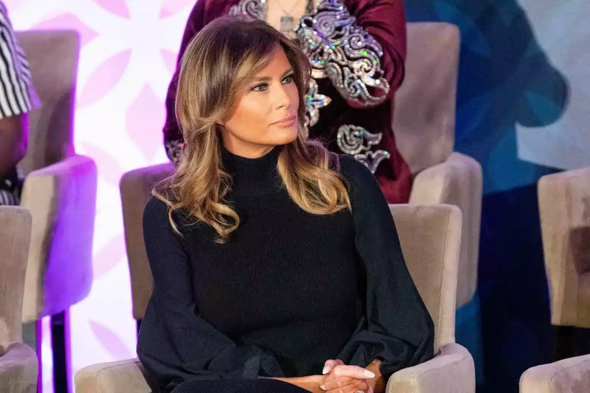 Melania Trump vestida amb un suèter negre de màniga llarga, està asseguda en una cadira beix, amb les mans entrellaçades sobre la falda, mentre mira cap a un costat en un entorn amb fons decoratiu i persones al voltant. Melania Trump vestida amb un suèter negre de màniga llarga, està asseguda en una cadira beix, amb les mans entrellaçades sobre la falda, mentre mira cap a un costat en un entorn amb fons decoratiu i persones al voltant.