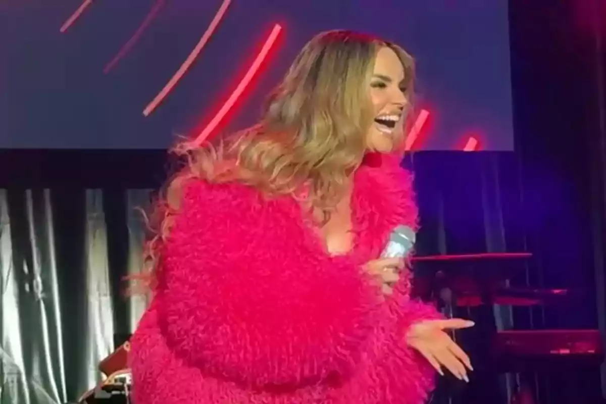 Melody amb abric rosa brillant somriu mentre sosté un micròfon en un escenari il·luminat Melody amb abric rosa brillant somriu mentre sosté un micròfon en un escenari il·luminat
