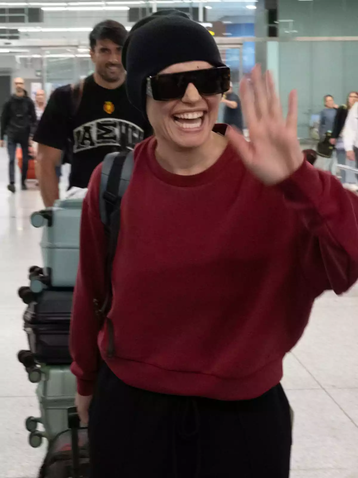 Melody somrient amb ulleres de sol i gorra negra saluda mentre camina per un aeroport, amb diverses maletes darrere. Melody somrient amb ulleres de sol i gorra negra saluda mentre camina per un aeroport, amb diverses maletes darrere.