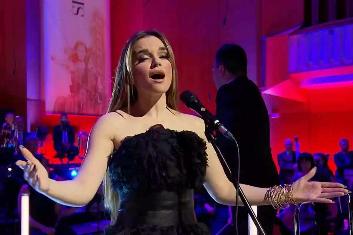 Melody canta apassionadament en un escenari il·luminat amb llums de colors, mentre el director d'Orquestra i Cor de RTVE es troba al fons. Melody canta apassionadament en un escenari il·luminat amb llums de colors, mentre el director d'Orquestra i Cor de RTVE es troba al fons.
