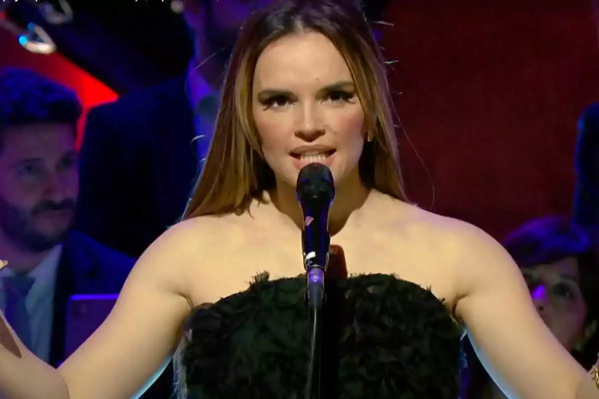 Melody canta en un escenari amb un micròfon acompanyada de l'Orquestra Simfònica de RTVE, vestida de negre, mentre el fons està il·luminat amb llums de colors. Melody canta en un escenari amb un micròfon acompanyada de l'Orquestra Simfònica de RTVE, vestida de negre, mentre el fons està il·luminat amb llums de colors.