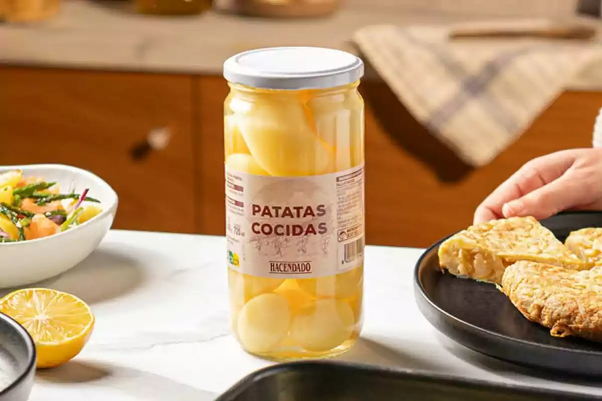 Un pot de patates cuites Hacendado sobre una taula al costat d'un plat de truita i una amanida.