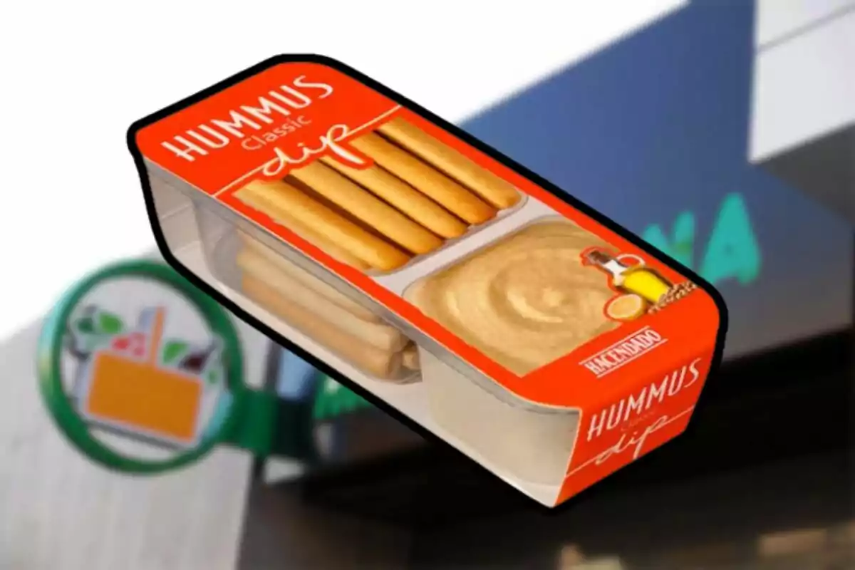 Envàs d'hummus clàssic amb bastonets de pa de la marca Hacendado davant d'un cartell de supermercat. Envàs d'hummus clàssic amb bastonets de pa de la marca Hacendado davant d'un cartell de supermercat.